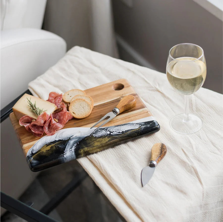 Wooden Cheese Charcuterie Serving Board with cured meats, bread, and cheese on a white blanket with a glass of white wine.