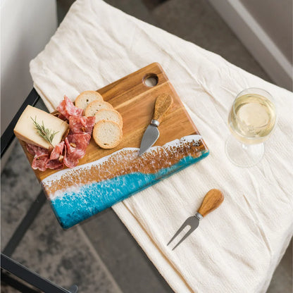 Wooden Cheese Charcuterie Serving Board with ocean design, bread, cured meats, and a glass of white wine on a white cloth.