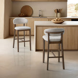Two mid-century modern bar stools with light gray upholstered seats and backs on solid wood legs, placed in a light wood kitchen with a smooth stone floor.