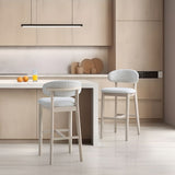 Two cream-colored upholstered bar stools with curved backs and light wood legs positioned beside a minimalist beige kitchen island with oranges and a glass of orange juice on top.