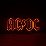 Red neon sign displaying the band name AC/DC with a lightning bolt symbol in place of the slash, set against a dark background.