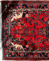 Corner of a hand-knotted Persian Hamadan wool rug with vibrant red field, intricate floral and geometric patterns in navy, beige, and green, framed by multi-layered borders.