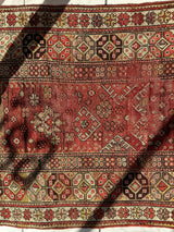 Close-up of an antique Turkish wool rug with distressed red background, geometric and floral patterns in cream, brown, and black, showing shadows cast across its surface.