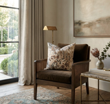 Brown armchair with woven wood sides holding a floral-patterned pillow featuring leaves in muted beige, gray, and brown, set beside a marble-top table and soft curtains.