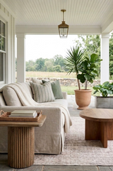 Outdoor porch seating with beige cushioned sofa, green and striped pillows, wooden round coffee table, potted plants, and a hanging lantern light under white paneled ceiling