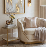 Cream-colored tufted armchair with brass nailhead trim, adorned with a pink antelope patterned pillow and beige throw, next to a gold and marble side table.