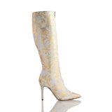 Tall white boot with gold floral damask pattern and a slim stiletto heel adorned with small metallic studs, shown from the side on a reflective surface.