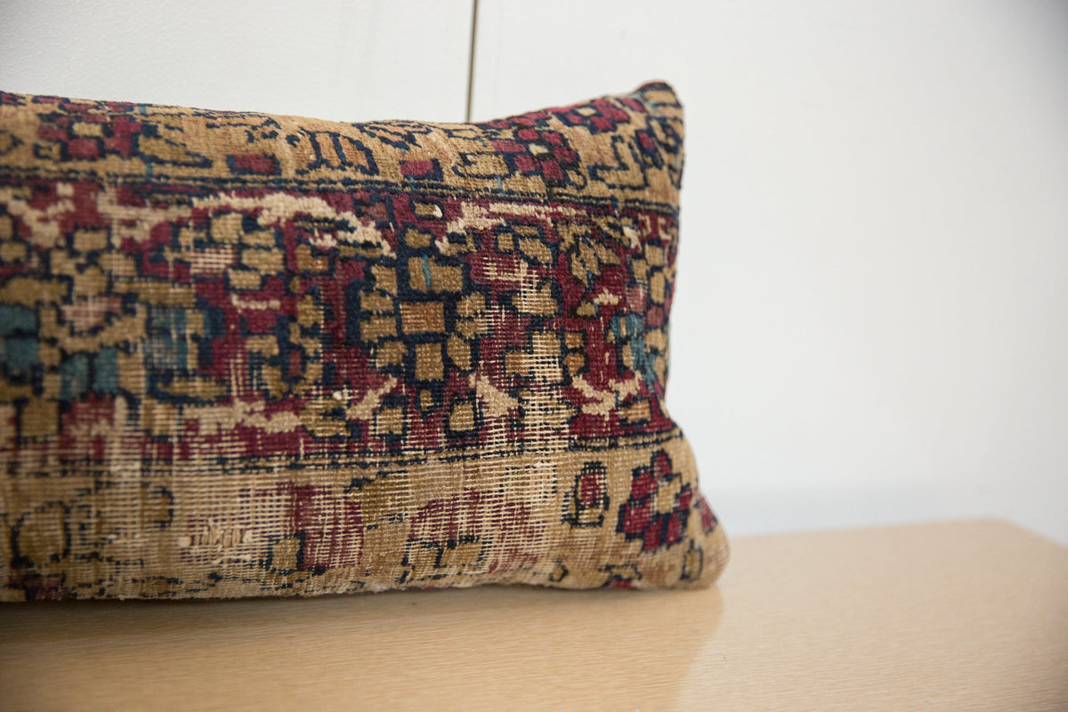 Close-up of a rectangular vintage rug fragment pillow with a worn beige background, intricate red and dark blue geometric patterns, resting on a light wood surface against a white wall.