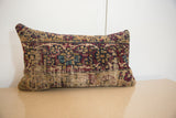 Rectangular pillow featuring a vintage rug fragment with faded red, blue, and beige geometric patterns set against a plain white wall and light wood surface.