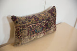 Rectangular pillow made from a vintage rug fragment featuring intricate patterns in burgundy, beige, and dark teal colors, placed on a light beige surface against a white wall.