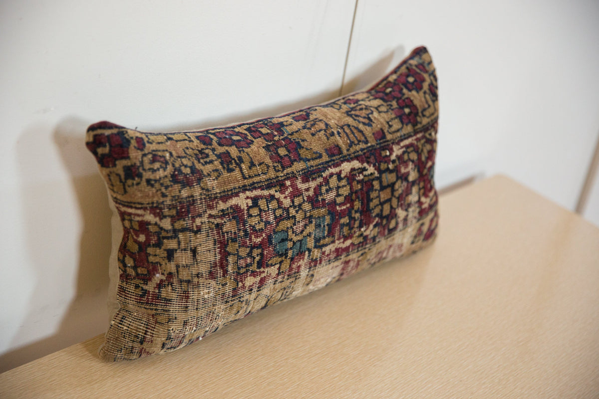 Rectangular pillow made from a vintage rug fragment featuring intricate patterns in burgundy, beige, and dark teal colors, placed on a light beige surface against a white wall.