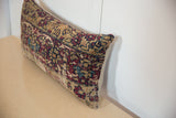 Rectangular pillow featuring a vintage rug fragment pattern with red, blue, and beige geometric motifs, resting on a light beige surface against a white wall.