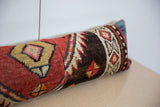 Long lumbar pillow covered in a vintage rug fragment with red, black, beige, and blue geometric patterns, resting on a light wooden surface against a white wall.