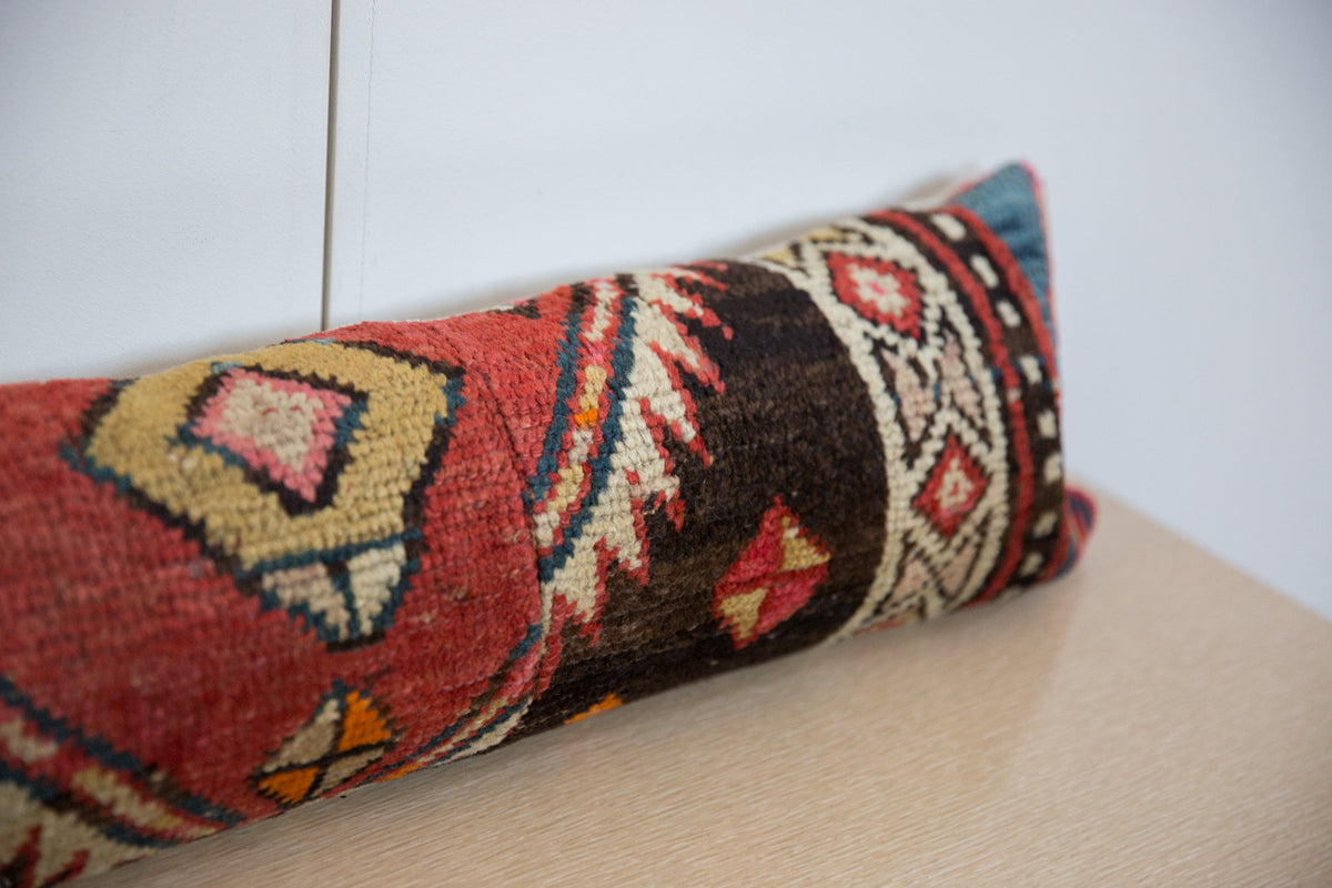 Long lumbar pillow covered in a vintage rug fragment with red, black, beige, and blue geometric patterns, resting on a light wooden surface against a white wall.