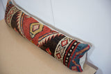 Long lumbar pillow featuring a vintage rug fragment with red, black, blue, and beige geometric patterns, resting on a light wooden surface against a white wall.