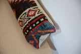 Rectangular lumbar pillow with a vintage rug fragment design featuring red, blue, black, and cream geometric patterns and a solid beige back, resting on a light wood surface.