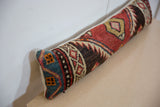 Long lumbar pillow with vintage rug fragment fabric featuring geometric patterns in red, blue, brown, and beige tones resting on a light beige surface.
