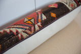Close-up of a lumbar pillow showing a colorful vintage rug fragment with red, orange, and beige geometric patterns on a dark brown background, contrasting with a plain light beige fabric.