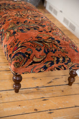 Rectangular ottoman upholstered with a vintage Persian rug featuring floral patterns in orange, navy, and olive tones, standing on turned wooden legs on a wood floor.