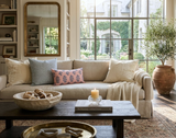 Neutral-toned living room sofa with blue, pink patterned Asha pillow cover, beige pillows, light throw, and rustic wooden coffee table with bowl and candle.