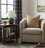 Beige boucle upholstered armchair with a textured sand-colored Audley Woven pillow, beside a dark wood side table holding a beige lamp and vintage airplane model, near a window with blue plaid curtains.