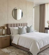 Modern bedroom with a taupe upholstered bed, white bedding, and two light blue floral pillows, complemented by beige walls and a round mirror above the headboard.