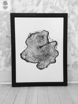 Black and white framed print of an Alaska Cedar wood slice showing detailed growth rings and irregular outer shape, leaning against a textured wall on a wooden floor.