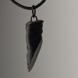 Small black pendant with textured surface and sharp angular design, hanging on a black cord against a gray background.