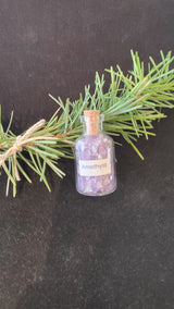 Small clear glass bottle labeled 'Amethyst' filled with tiny purple crystal chips, placed next to a green pine branch on a dark surface.