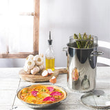 Tall stainless steel pot with a steamer basket holding asparagus, next to a pie with colorful beet slices on a white textured table by a wooden window, garlic, and olive oil bottle.