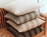 Stack of four square pillows on a striped wooden chair, featuring textured cream on top and three patterned covers below in black, brown, and beige tones with dotted designs.
