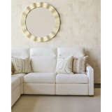 Cream sectional sofa with textured beige and patterned fawn brown pillows arranged on it, set against a light patterned wall with a round gold decorative mirror above.