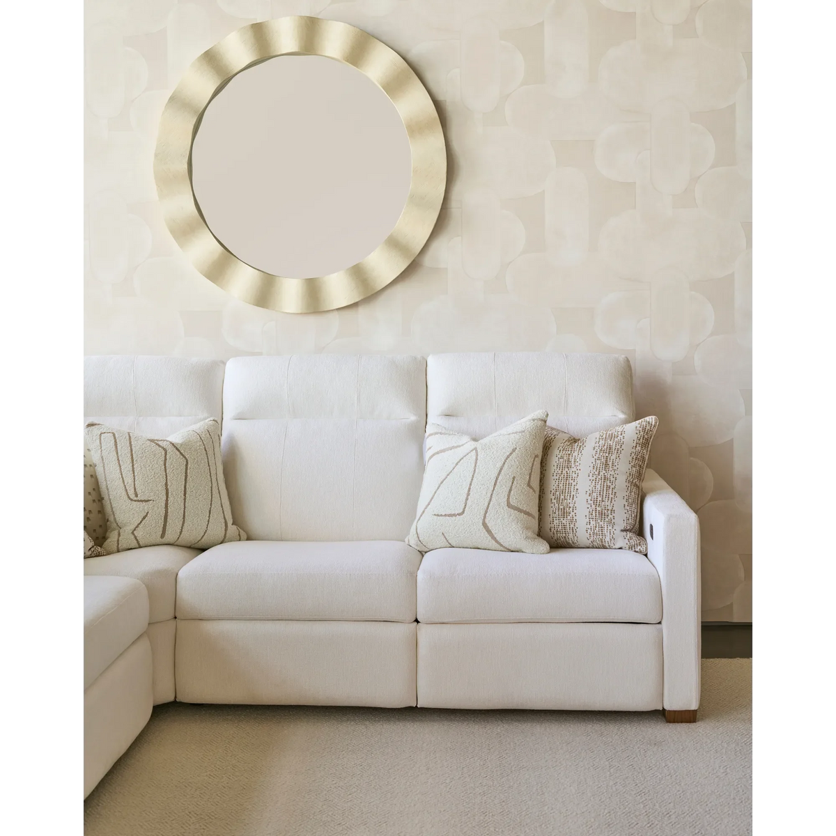 Cream sectional sofa with textured beige and patterned fawn brown pillows arranged on it, set against a light patterned wall with a round gold decorative mirror above.