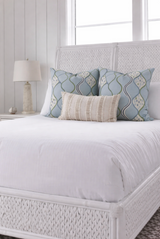 Bed with white woven headboard and footboard, featuring two blue patterned square pillows and one beige textured lumbar pillow on white bedding.