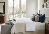 Neutral-toned bedroom with beige upholstered bed, white bedding, beige textured throw, and a mix of navy blue and patterned pillows, flanked by two wooden nightstands.