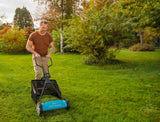 Man in brown shirt and beige pants pushing a blue cylinder lawnmower on a green grassy lawn surrounded by autumn-colored trees and shrubbery.