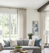 Light gray sofa with five pillows in cream, light blue, and bold blue-line pattern, positioned between two tall windows with cream curtains and wooden beam ceiling.