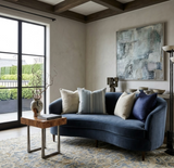 Curved navy blue velvet sofa with assorted neutral and blue pillows against a light gray wall, featuring abstract artwork and a wooden side table with sculptural decor.
