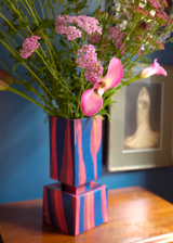 Squared vase composed of two stacked cubes with vertical blue and pink stripes, holding pink and green flowers, set on a wooden surface against a blue wall.