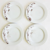 Four white dessert plates arranged in a square, each featuring a delicate hand-drawn brown birch tree design on one side with seasonal twig details.