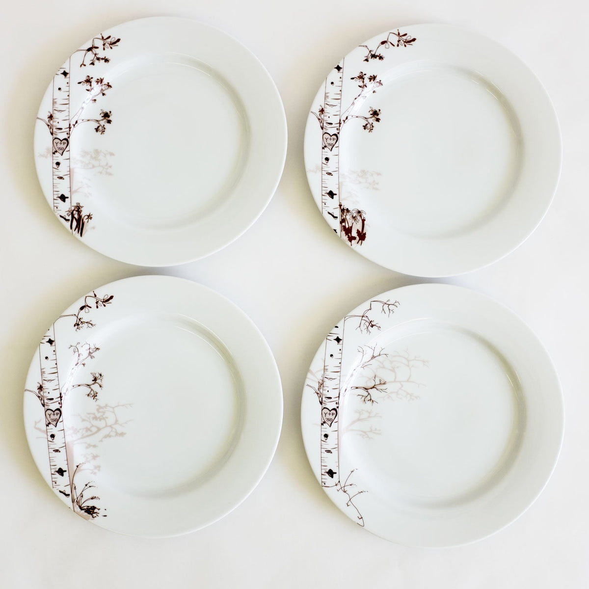 Four white dessert plates arranged in a square, each featuring a delicate hand-drawn brown birch tree design on one side with seasonal twig details.