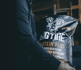 Person in dark clothing holding a large black bag labeled 'BIG TINE PROTEIN PLUS' featuring a white deer head logo and text about purified deer blend and soybeans.