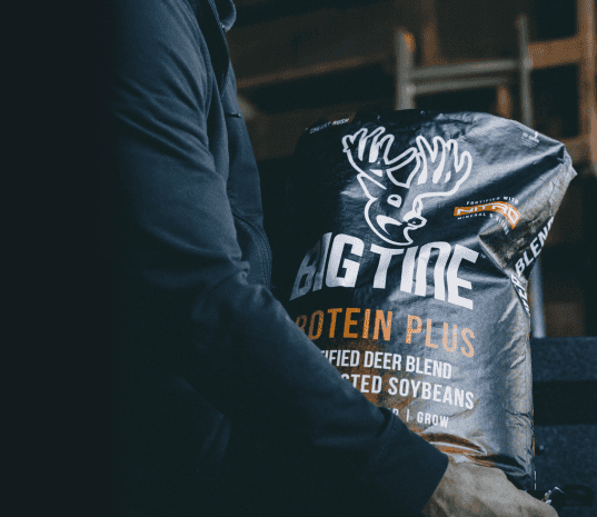 Person in dark clothing holding a large black bag labeled 'BIG TINE PROTEIN PLUS' featuring a white deer head logo and text about purified deer blend and soybeans.