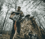 Man wearing outdoor clothing and cap holding a black bag labeled 'BIG TINE NITRO' in a leafless forest standing next to a weathered tree stump, viewed from low angle.
