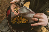 Hands holding a mix of yellow and black deer feed kernels above an open bag labeled Big Tine Fortified Deer Blend Cherry, outdoors in a natural setting.