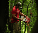 Man in a red shirt and beige pants pouring granular material from a red and white bag into a forested area with green leafy trees around.