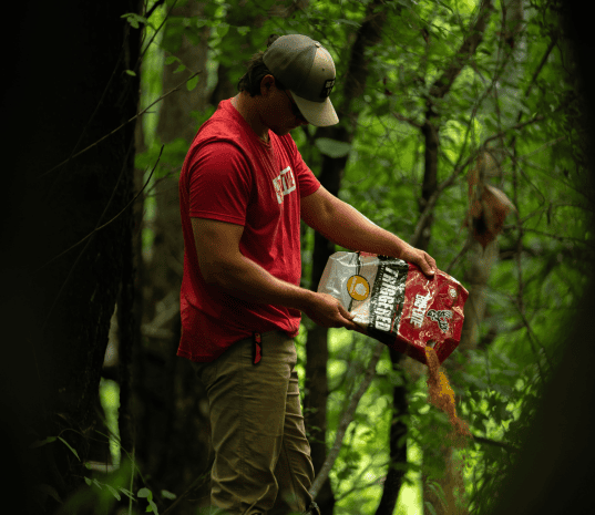 Man in a red shirt and beige pants pouring granular material from a red and white bag into a forested area with green leafy trees around.