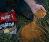 Hand pouring loose orange powder onto green grass with a red and white Big Tine Triggered Pear branded bag nearby outdoors.