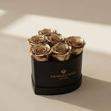 Six gold-colored roses arranged in a black heart-shaped box with gold lettering on a beige surface with soft shadows