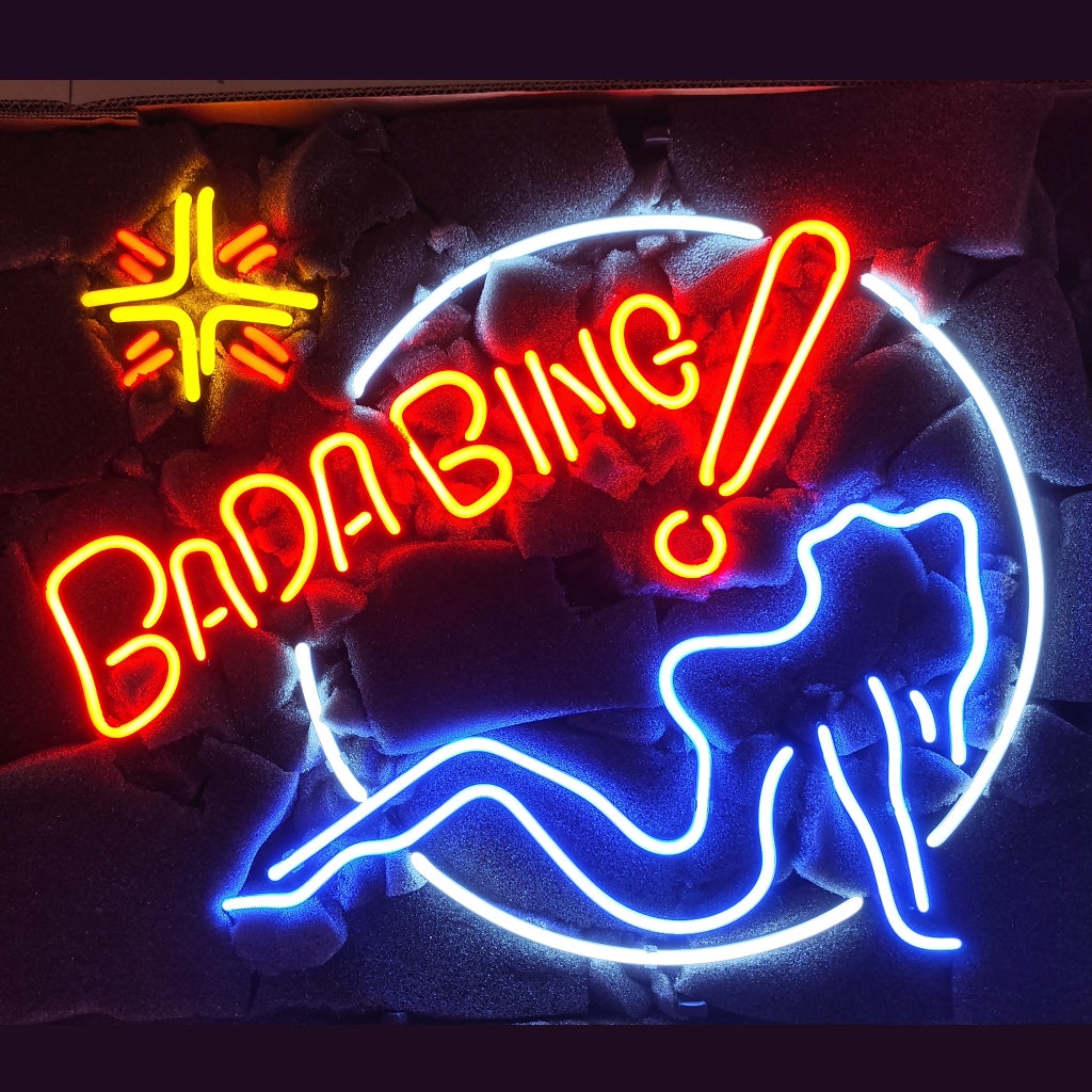 Neon sign with the text 'BADA BING!' in red, a stylized reclining woman outline in blue, and a yellow starburst shape on a black textured background.